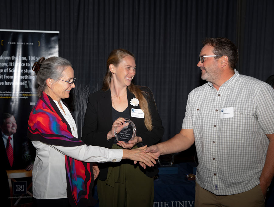 A woman and a man shake hands in front of another woman holding a glass award.