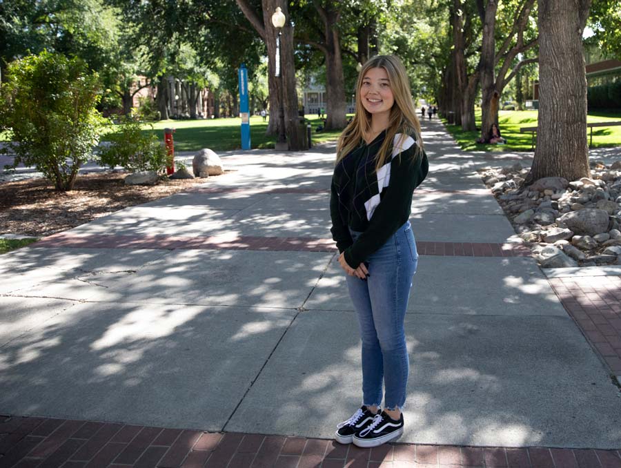 A girl with blonde hair stands along a pathway lined with tall oak trees. She is smiling and wearing jeans and a black sweater.