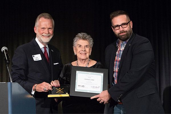 KNPB CEO and emcee Kurt Mische poses next to Nevada Writers Hall of fame founder Marilyn Melton as they present Caleb S. Cage with the 2019 Silver Pen Award. Caleb smiles, holding his plaque.