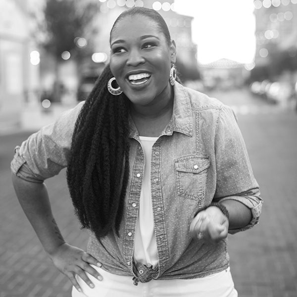 Portrait of Vogue Robinson wearing white pants and a white tank top with a denim button-up shirt on top. She has one hand on her hip and is gesturing with the other hand. She is displaying a warm smile.