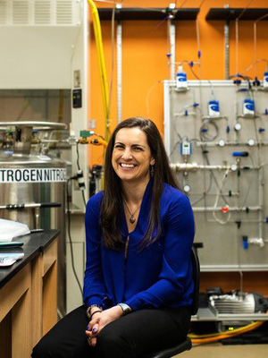 Sarah Horst smiles, sitting at a lab bench with equipment around her, including a large canister of nitrogen.