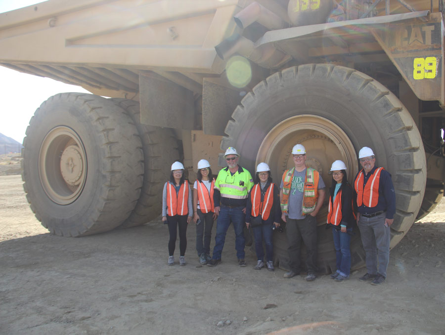 A group of seven people wearing orange high-visibility vests and white hard hats stand next to a massive mining vehicle tire.