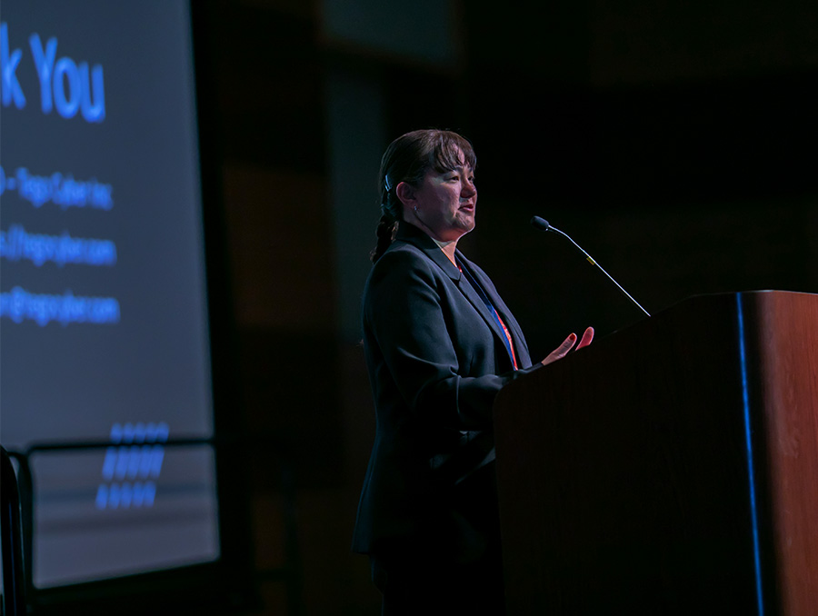 Profile of a woman facing right, standing behind a podium, section of a projection screen behind her.