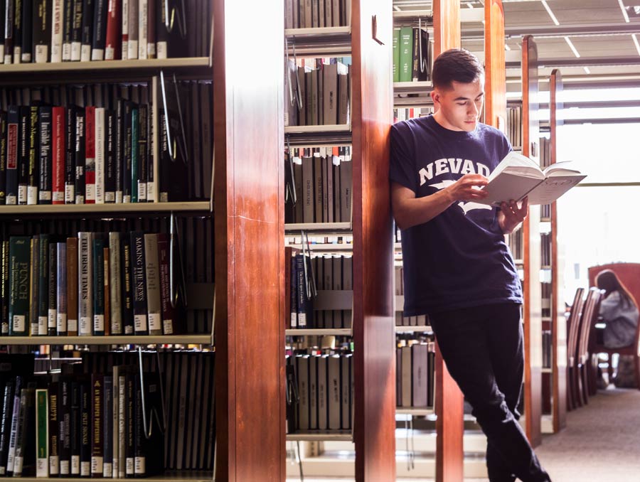 A student wearing a Nevada t-shirt leans against a bookshelf in the Knowledge Center with a book in his hands, reading.