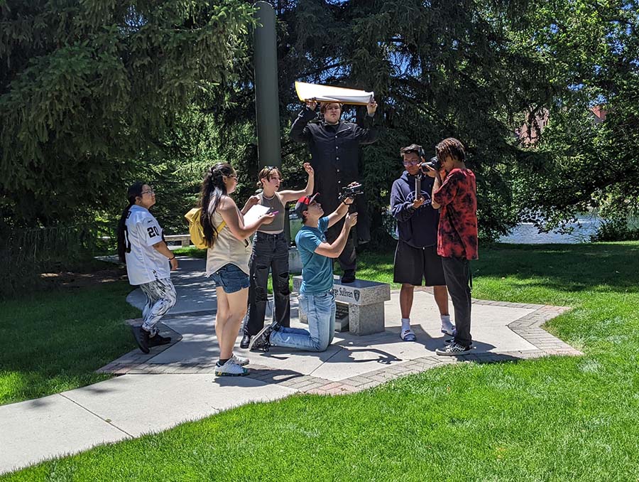 Group of seven students shoot a scene for their documentary-style film. Students are seen holding cameras, screens, microphones and video camera.