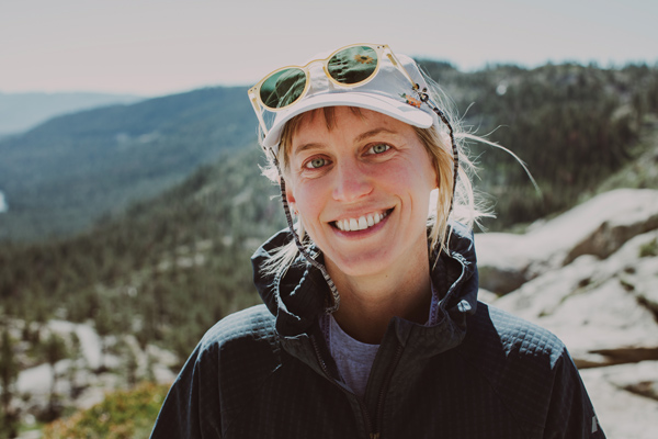 A woman wearing a hat smiles, looking at the camera. There are mountains and rocks in the background.