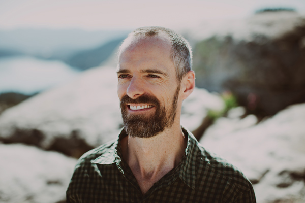 A man looks away from the camera, smiling. He is in front of a rocky, mountainous terrain.