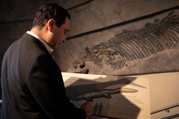 A man wearing a black suit stands in front of a display featuring an ichthyosaur fossil, reading the description.