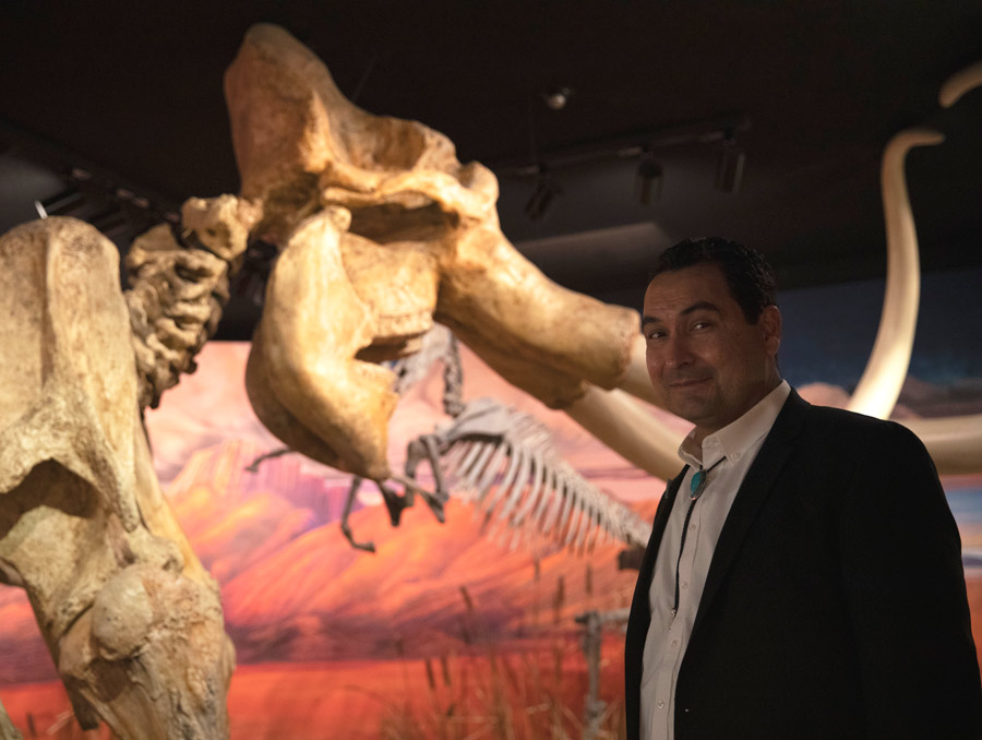 A man in a black suit and turquoise bolo tie smiles in front of a cast of a mammoth skeleton.