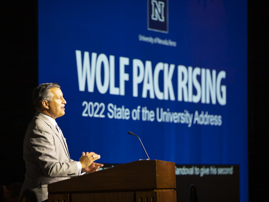 UNR President Brian Sandoval stands at the podium giving the 2022 State of the University address with a slide titled Wolf Pack Rising behind him.