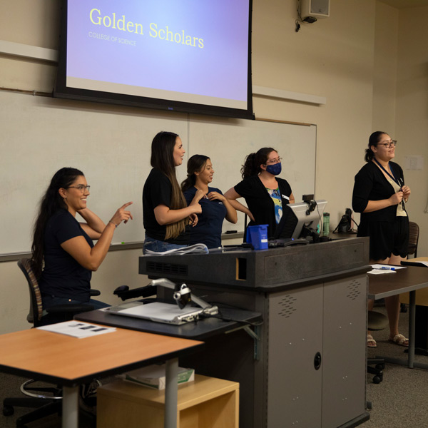 Five women stand at the front of a classroom in front of a projector screen that reads "Golden Scholars."