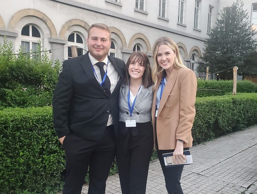 Three students - one boy and two girls (from left to right) - dressed in professional attire standing in front of a building and some bushes. 