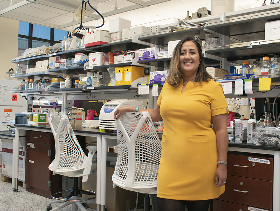 Assistant Professor Maryam Sarmazdeh stands in a yellow dress before a wall of shelving filled with equipment in her lab.