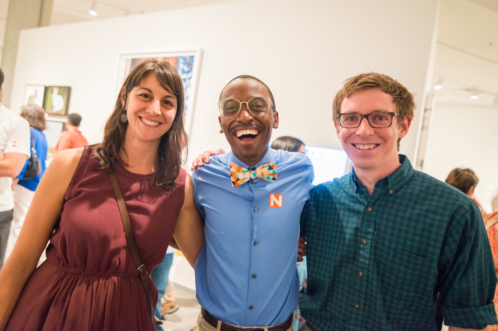 Three students, a woman and two men, post in nice clothing smiling at the camera while wearing a Nevada Museum of Art admittance sticker.