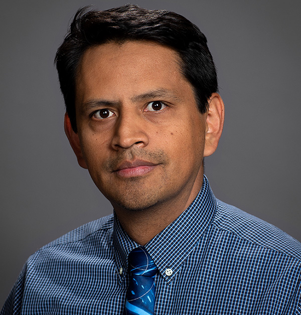 Portrait of Carlos wearing a dark blue, long-sleeve button-up shirt and blue necktie.