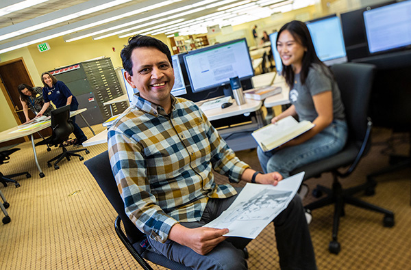 Carlos sits in a chair in front of a table with computers with a female student. They are each holding a book and smiling.