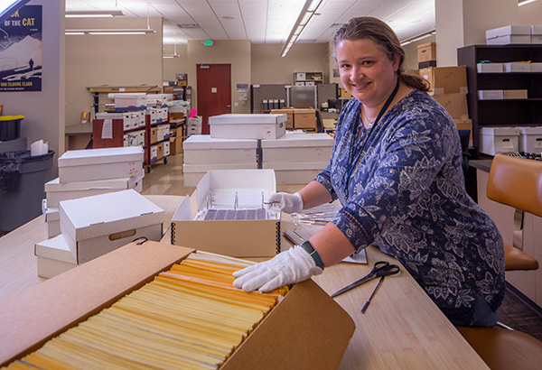 Elspeth stands at a work table with several boxes of photo negatives. She is rehousing original photo negatives from their original envelopes into new, archival-quality storage sleeves and binders.