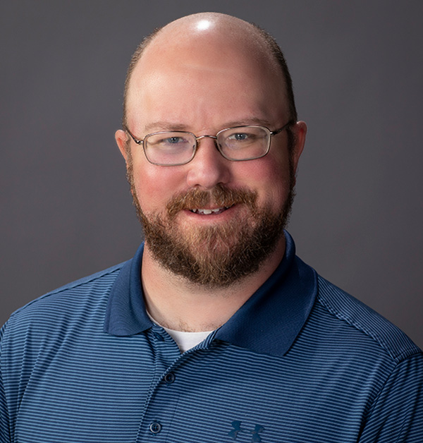 Portrait of Mike wearing wire-framed glasses, a dark blue shirt and a white undershirt.