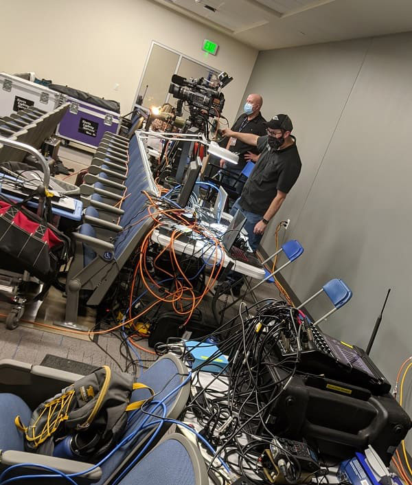 Two men stand behind a table full of laptop computers, video camera and sound equipment, as well as several sets of wires and extension cords. Shawn Sariti operating the camera and Kyle Weerheim monitoring equipment during one of the University's many virtual events.