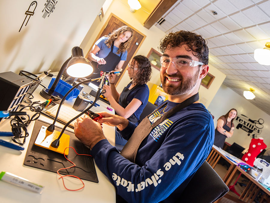 Two people sit at a desk equipped with soldering equipment and supplies. They each are using the equipment to make something. There are other people working and hanging out in the background.