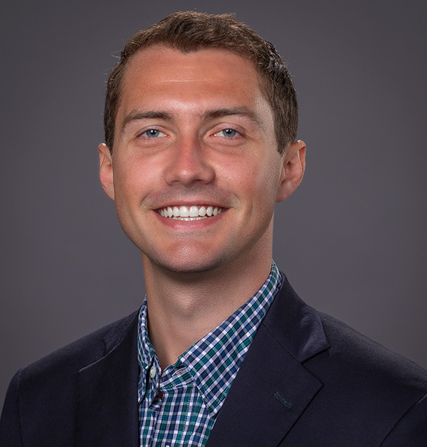 Portrait of Teddy wearing a dark sport coat with a blue and white, checkered-pattern collared shirt. 