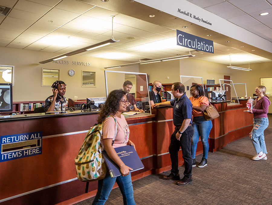 Four University Libraries users check out and drop off materials at the Access Services desk located on the second floor of the Mathewson-IGT Knowledge Center. There are three student assistants working behind the Access Services desk. One is taking a phone call, one is stamping a due date inside the back of a book being checked out and the third is working on a computer.