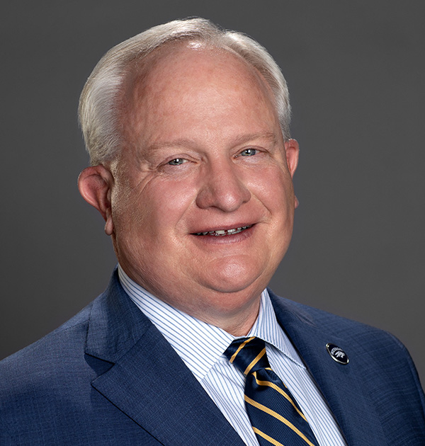 Portrait of Provost Jeff Thompson wearing a navy blue suit with a white and light blue striped collared shirt and a navy blue tie with yellow stripes.