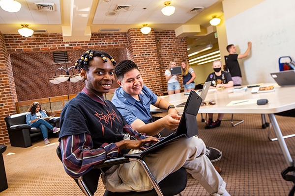 Group of seven students study inside the DeLaMare Science & Engineering Library. They are using the white board on the wall to take notes, are seated around a table with laptops and tablets.