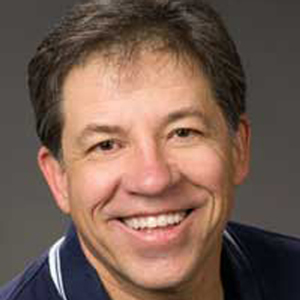 A man smiles, wearing a blue polo shirt in front of a gray background.