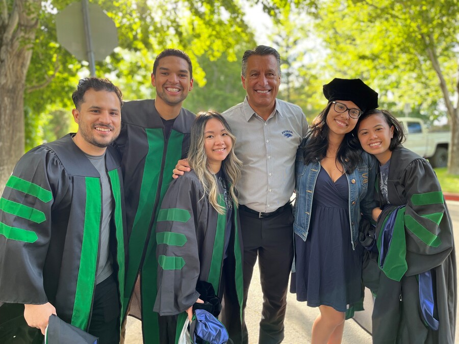 University President Brian Sandoval poses outside on the campus quad with a group of students who are all dressed in Med School regalia.