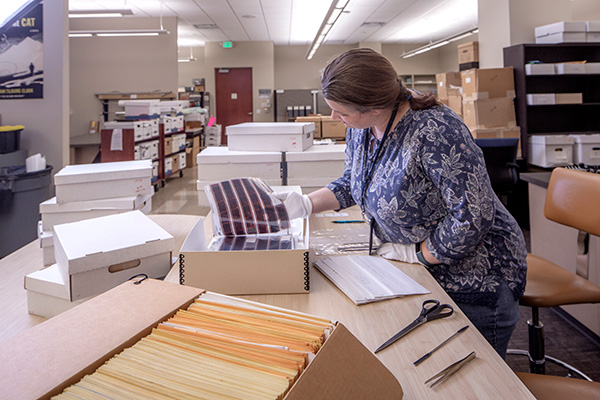 Elspeth standing behind table with many boxes visible. One box is in the foreground containing hundreds of envelopes filled with numerous photographic negatives. Tools used to process the collection can also be seen. 