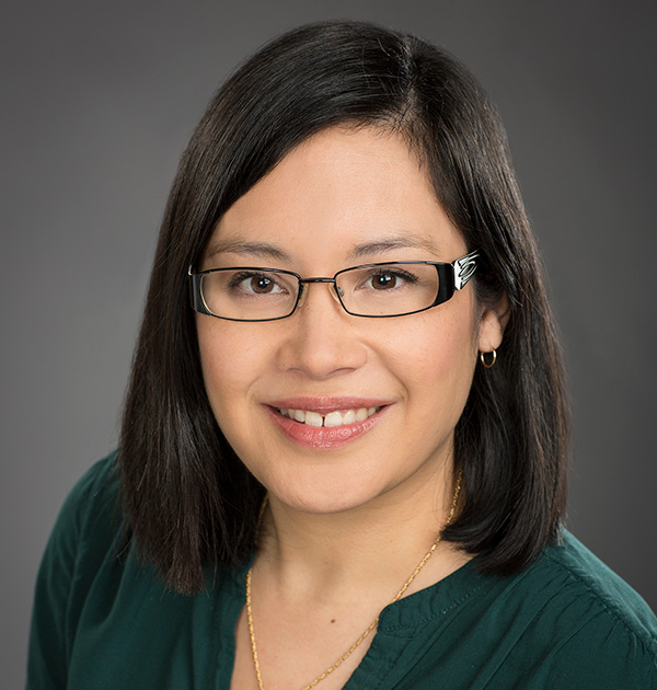 Portrait of Katherine Dirk wearing dark, wire-frame glasses, a gold necklace and an emerald green blouse.