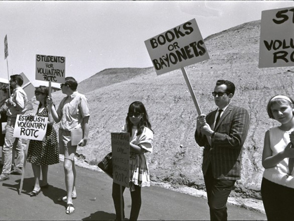 A group of seven men and women dressed in suits, dresses and full hair and make-up holding protest signs that read “Books or Bayonets,” “Students for Voluntary ROTC,” and “Establish Voluntary ROTC”