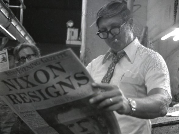 Man with dark-rimmed reading glasses wearing a white short sleeved shirt and tie with a wedding band and gold watch holds a copy of the Reno Gazette Journal newspaper where the front page can be seen. It says “Nixon Resigns.”