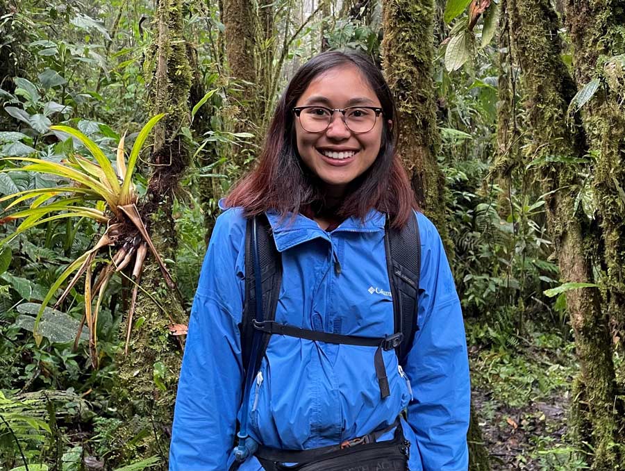 Chanchanok Sudta wearing a bright blue jacket stands in a jungle surrounded by foliage, smiling.