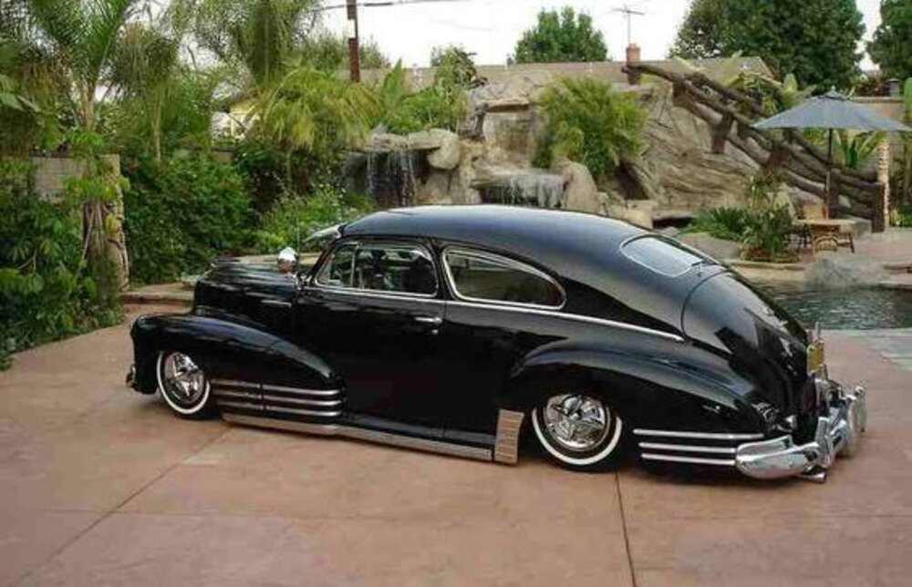 A vintage looking black lowrider car parked by a beautiful water feature or fountain.