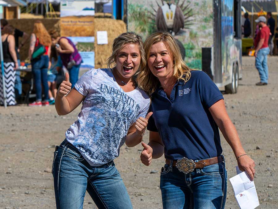 Two smiling University students at Nevada Field Day in Wolf Pack gear pose with thumbs up for a photo. Behind them are hay bales and people of all ages at info booths and activity trailers.