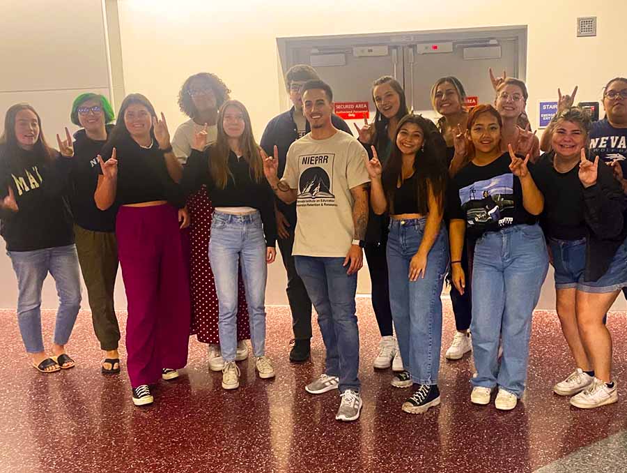 A group of about 14 Nevada Institute on Educator Preparation fellows at the airport pose holding up the Nevada Wolf Pack hand signal.