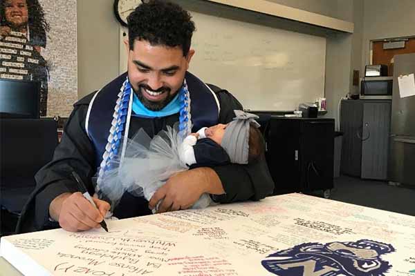 Ortiz with graduation gown, holding baby and signing poster