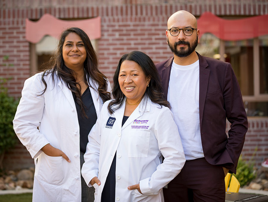 Two people in white lab coats and a person in a sweater pose in front of a brick building.