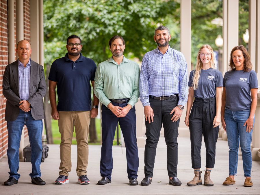 Four men and two women standing on the porch of Morrill Hall smiling for group photo.