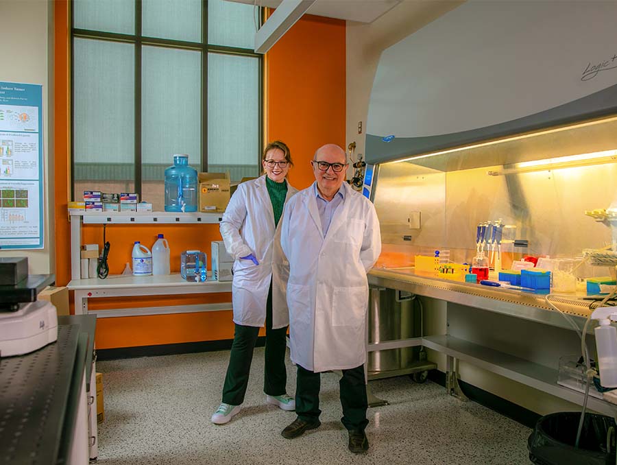 Two people, a younger woman and older man, wearing white lab coats and standing in a lab. 