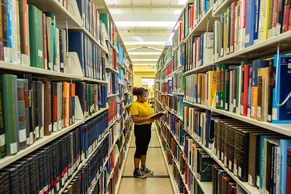 A university students stands between two stacks of books. She is holding a book and is reading it while standing in the stacks.