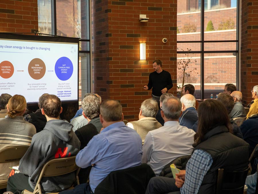 A man in a black long-sleeved t-shirt presents at the front of a room full of people. He is pointing a clicker at a screen.