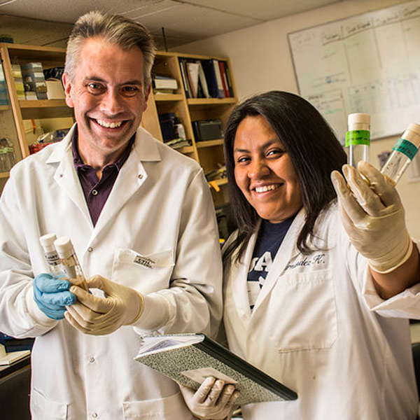 Tom Kidd and Karla Hernandez in the lab, smiling and wearing lab coats. They are both holding tubes of fruit flies.