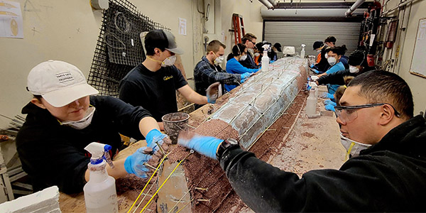 Students wearing masks and blue gloves applying concrete to a canoe mold.