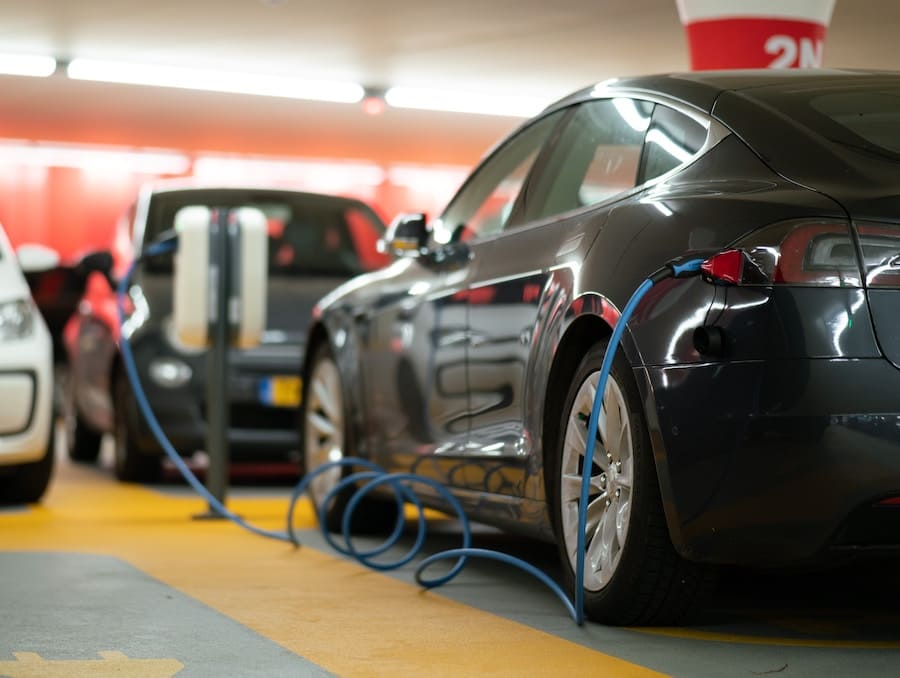 Dark gray Tesla parked in a public garage next to wide yellow stripe on the ground, with a blue cord plugged into its charging port.