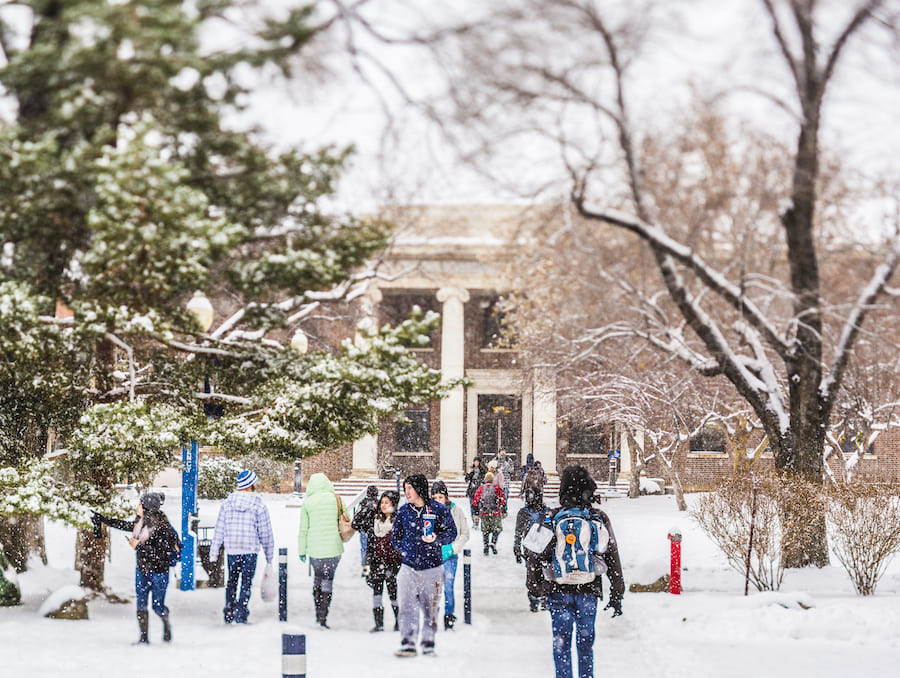 Students walking on a path covered in snow surrounded by trees covered in snow and a brown building with white columns in the middle.