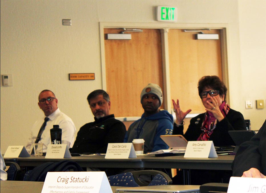 Four people sitting inside at a long table; the person on the right is speaking and gesturing with hands in front of her body.