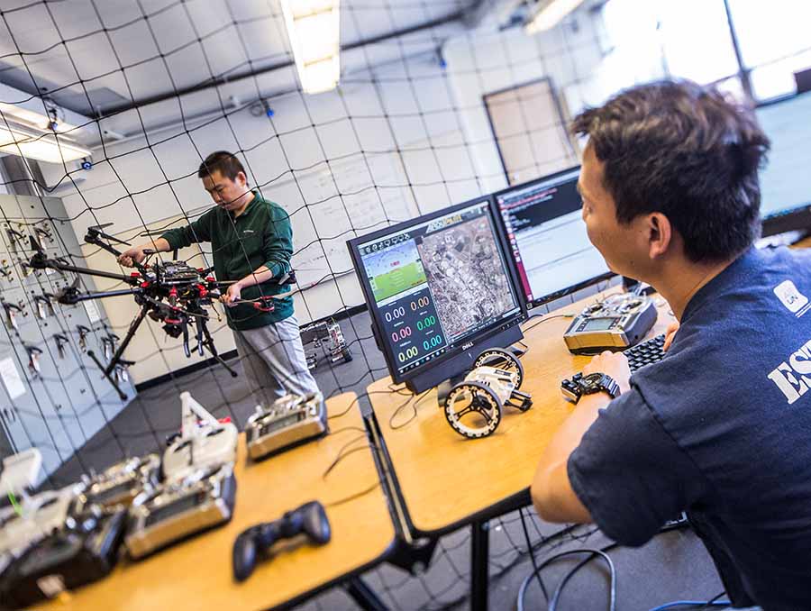 Two students standing in a lab; the student in the background is holding a drone and looking at it; the student in the foreground  is a a computer monitor and also is looking at the drone.